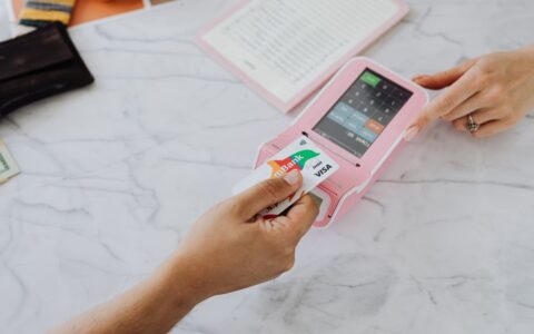 Hands making a contactless payment using a credit card and a pink terminal on a marble table.