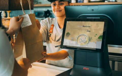A smiling cashier hands a paper bag to a customer at a modern point of sale, showcasing customer service.