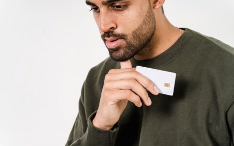 Pensive man in green sweater holds a white credit card against a white background.