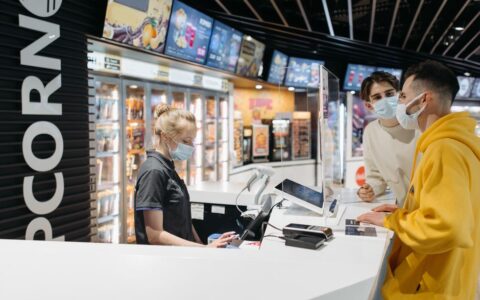 Customers wearing masks interact with a cashier at a cinema concession stand, following safety protocols.