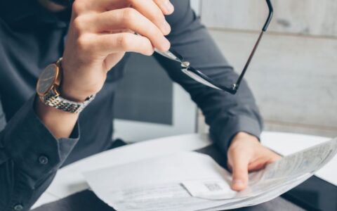 Man in formal attire reviewing paperwork, holding glasses. Business setting.