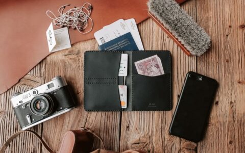 Flat lay of traveler essentials including wallet, camera, and phone on wooden surface.